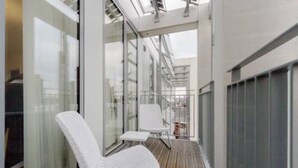 Un balcon avec une chaise blanche et une petite table, des murs en verre et une vue sur le paysage urbain.