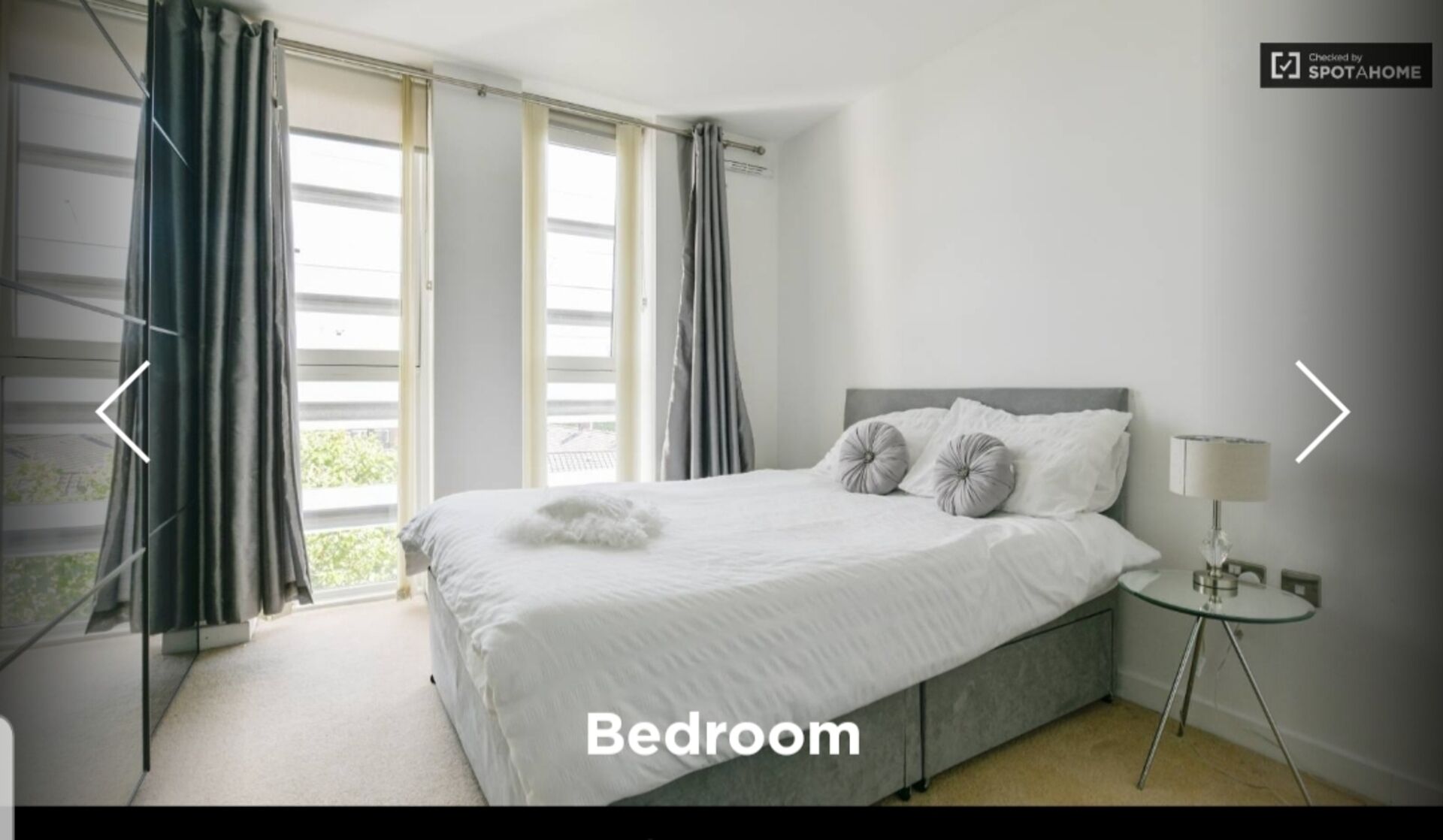 A bedroom with a bed, a bedside table with a lamp, and a window with curtains.
