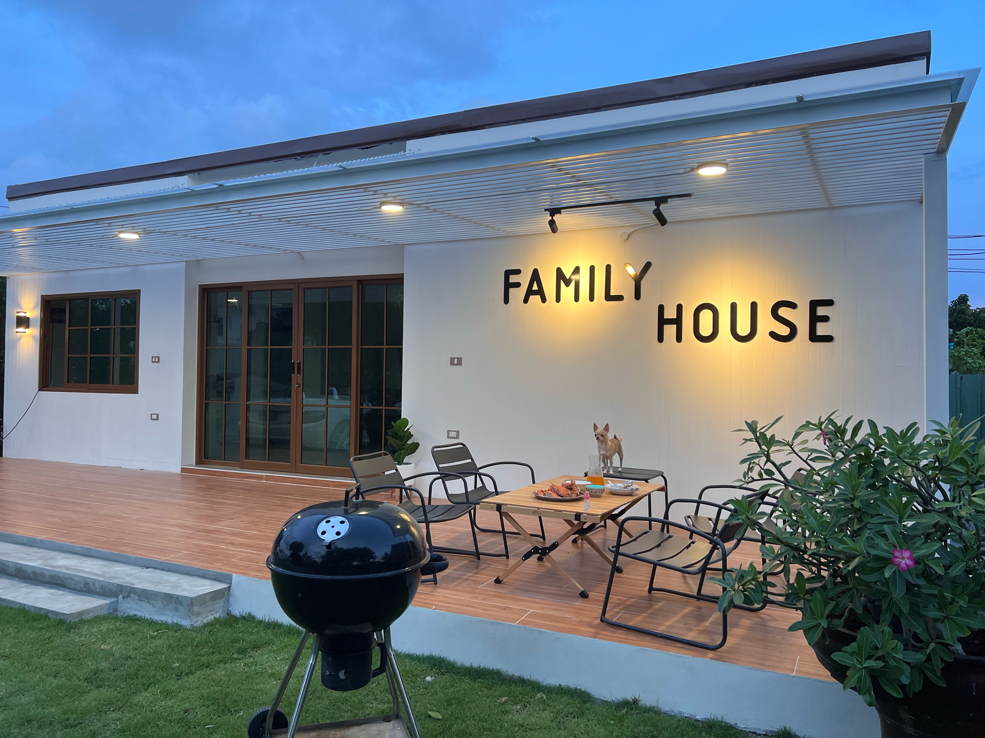 Una casa moderna con un letrero que dice 'FAMILY HOUSE', una parrilla, mobiliario de exterior y una puerta de cristal de entrada.