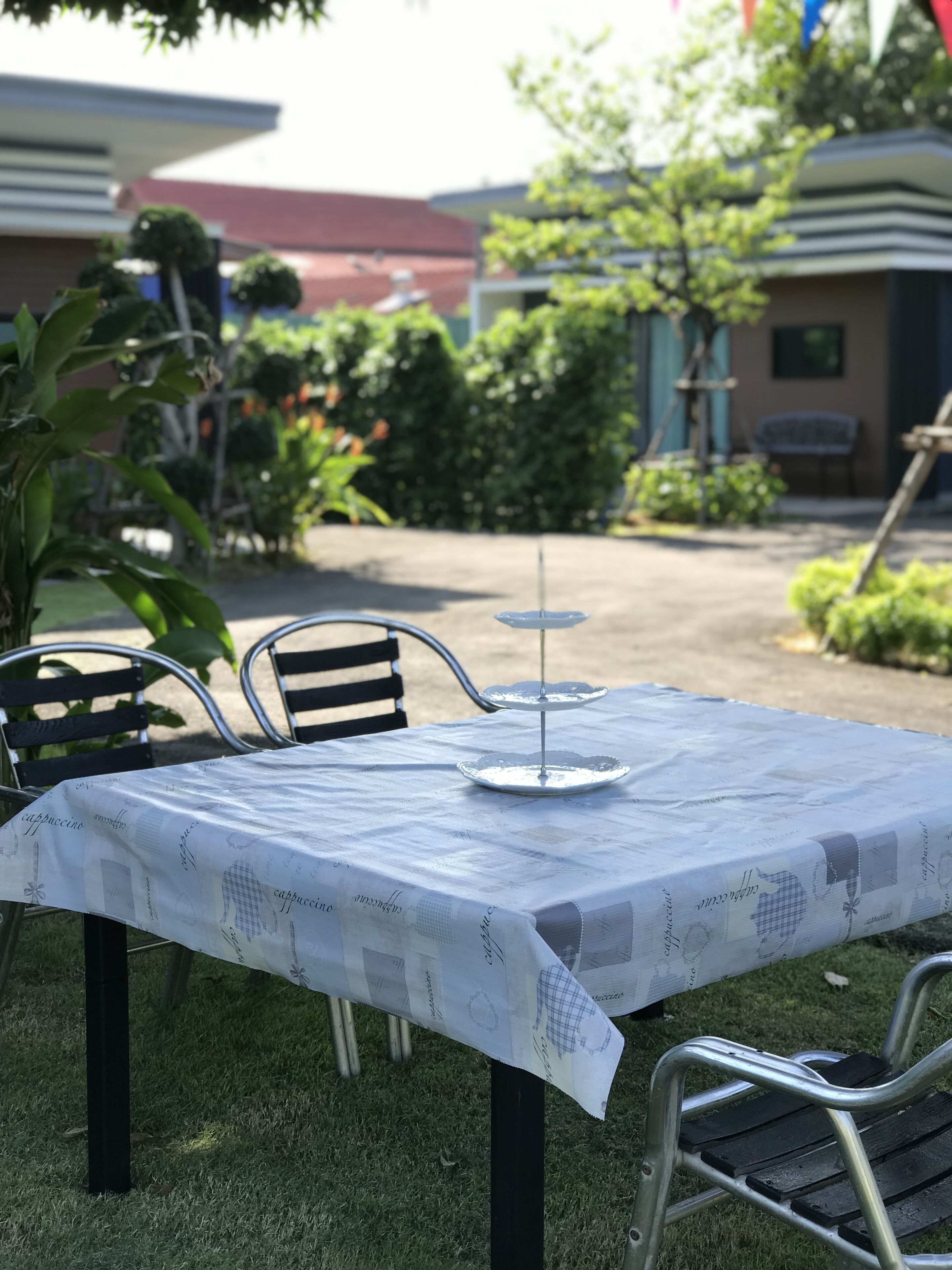 An outdoor table with a white tablecloth and metal chairs, set in a grassy area with buildings and trees in the background.