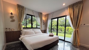 A modern bedroom with a large bed, a nightstand, a lamp, and a view of greenery through large windows. - Bannjamjuree Resort Sattahip (Sattahip)