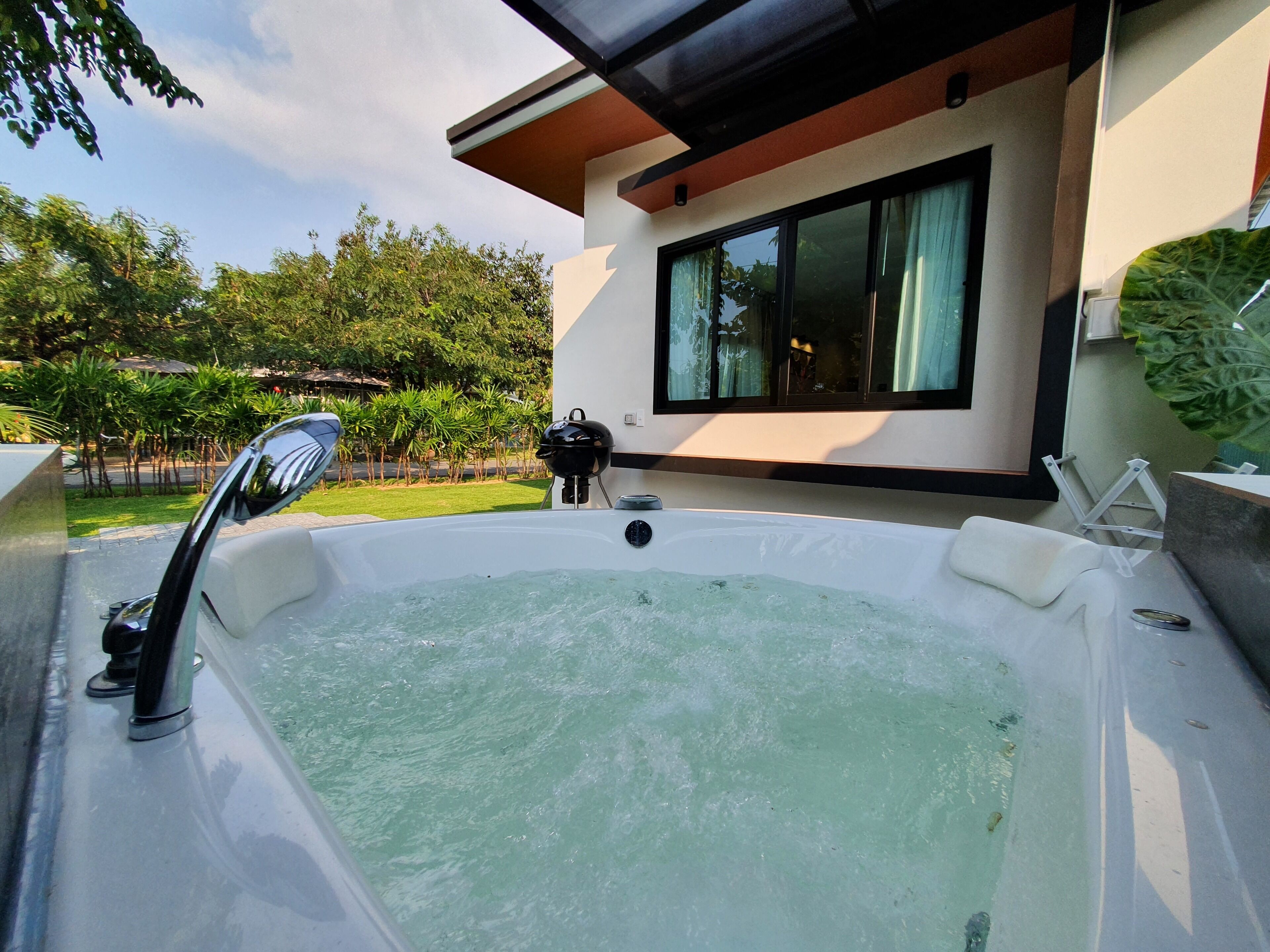 Un jacuzzi con una parrilla negra, una ventana con cortinas y vistas a un jardín con palmeras.