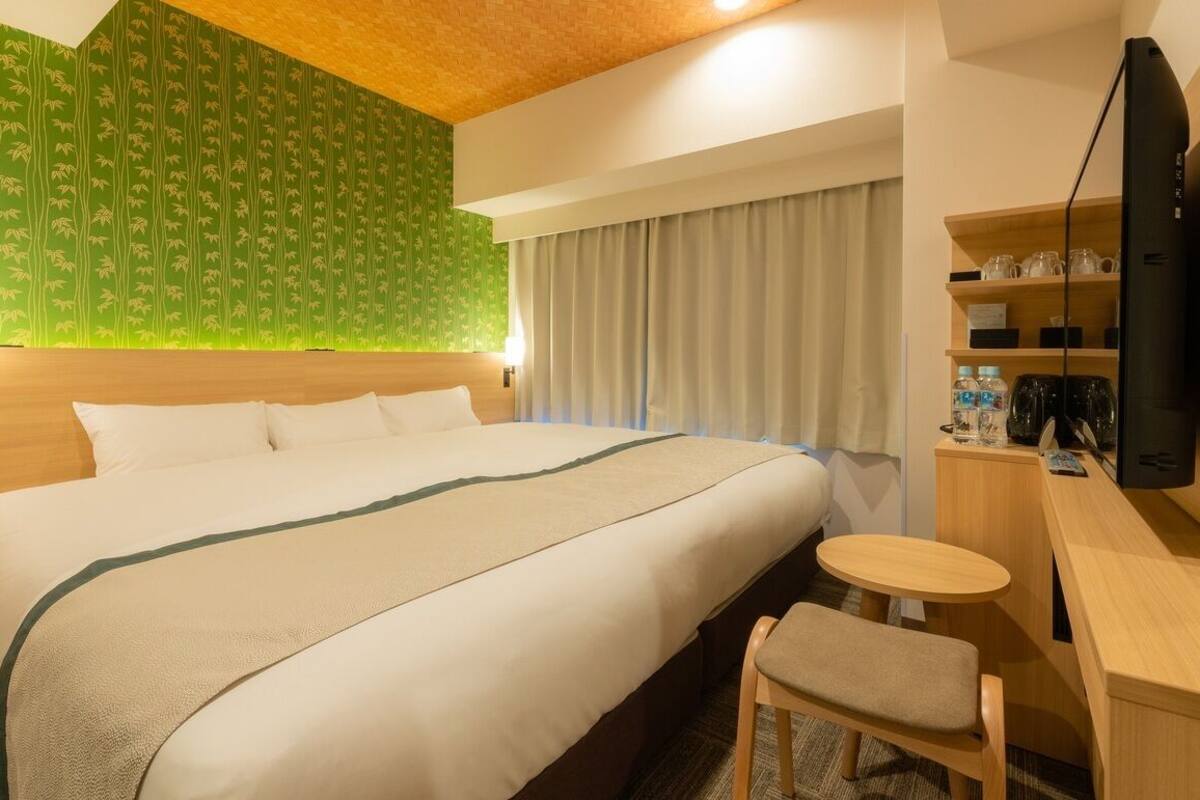 A hotel room with a large bed, a desk with a chair, a television, and a shelf with items.
