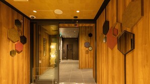 A modern hotel lobby with wooden paneling, a glass elevator entrance, and geometric wall decorations.