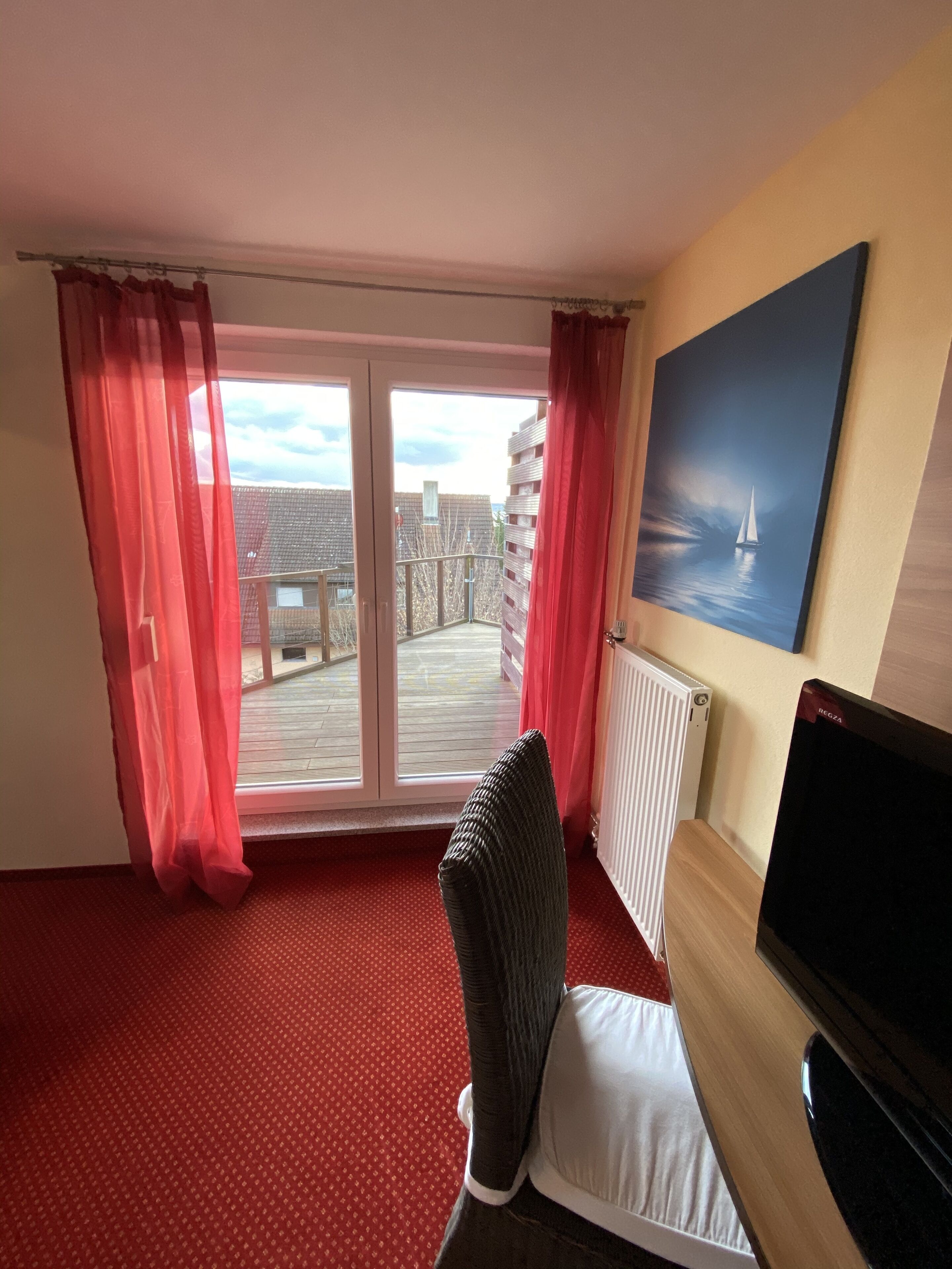 A hotel room with a red carpet, a chair, a TV, a painting of a sailboat, and a view of a balcony with buildings.