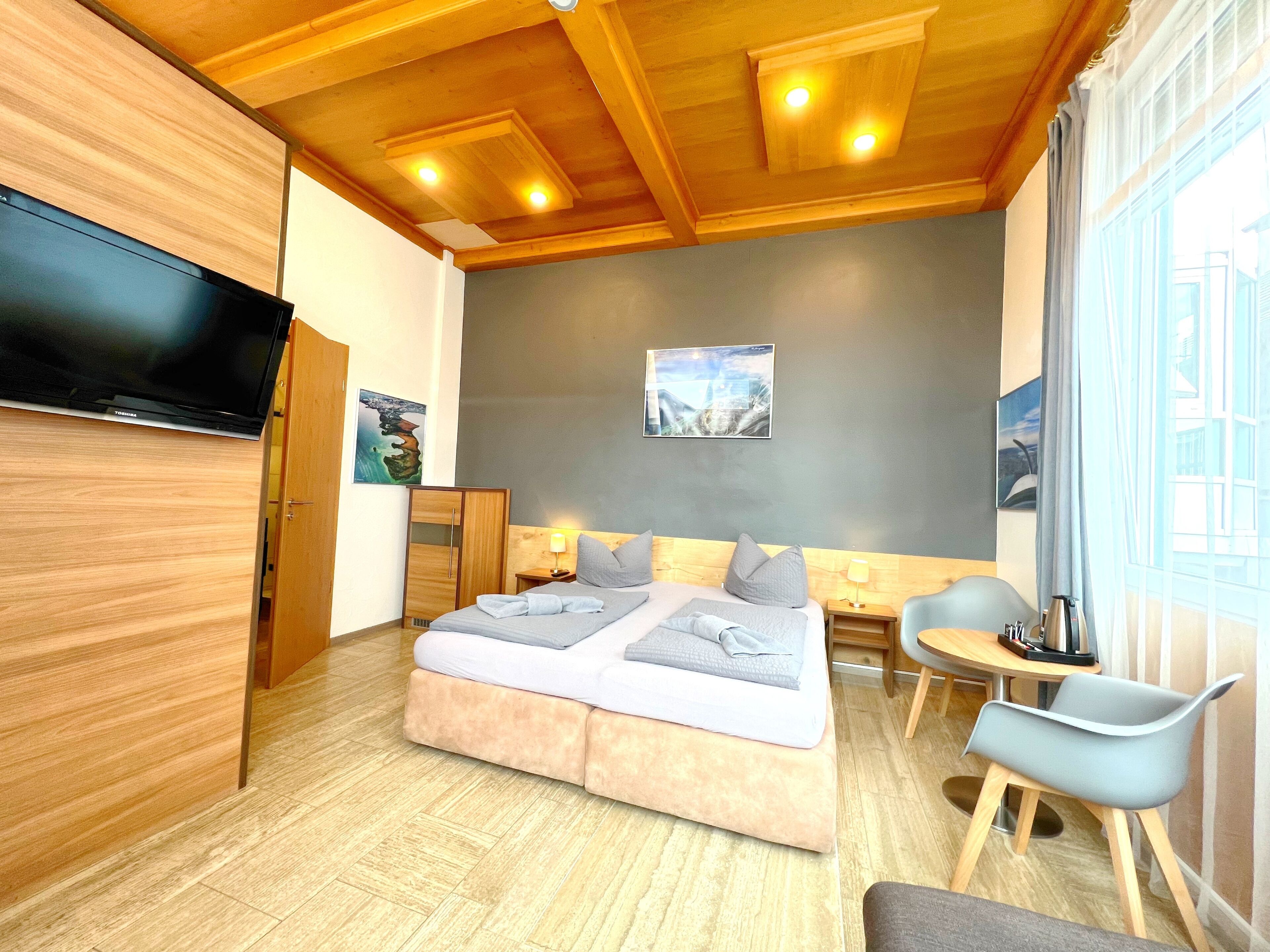 Habitación de hotel moderna con una cama grande, televisor de pantalla plana, techo de madera, obras de arte y un área de comedor con una silla.