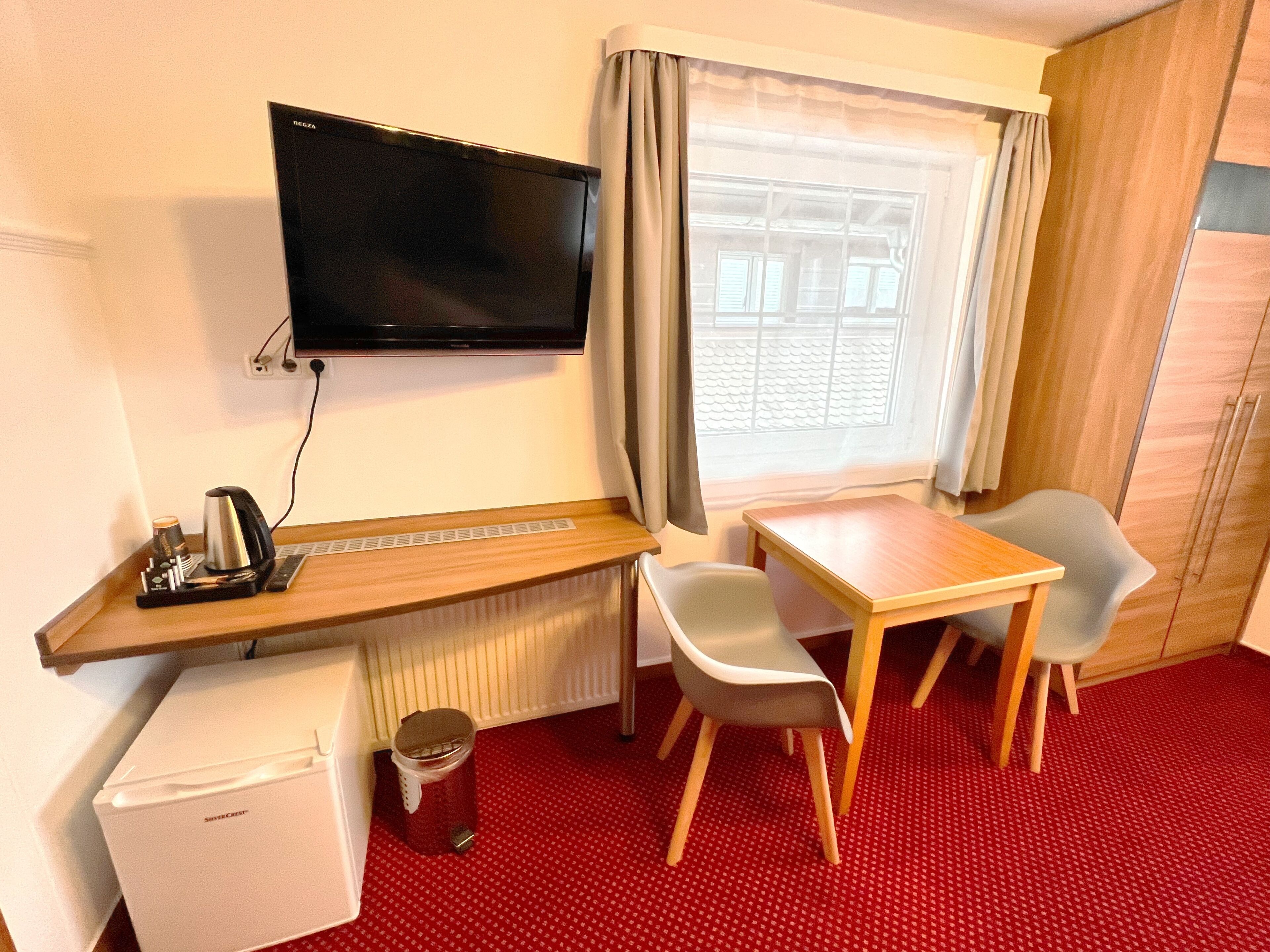 A hotel room with a television mounted on the wall, a small refrigerator, a table with two chairs, and a window with curtains.