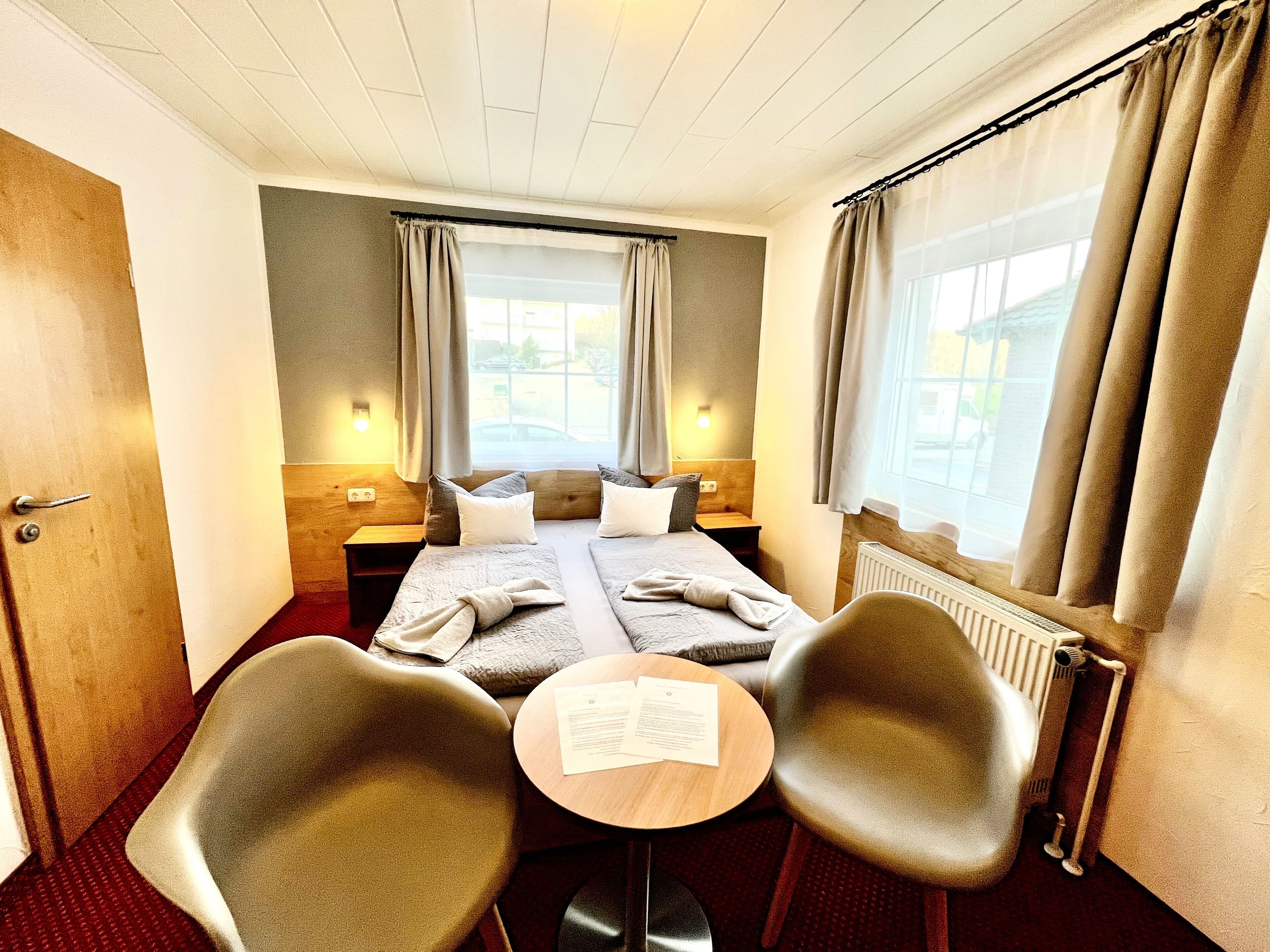A hotel room with two beds, a small table, two chairs, a window with curtains, and a radiator.