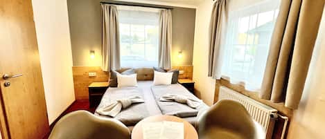 A hotel room with two beds, a small table, two chairs, a window with curtains, and a radiator.