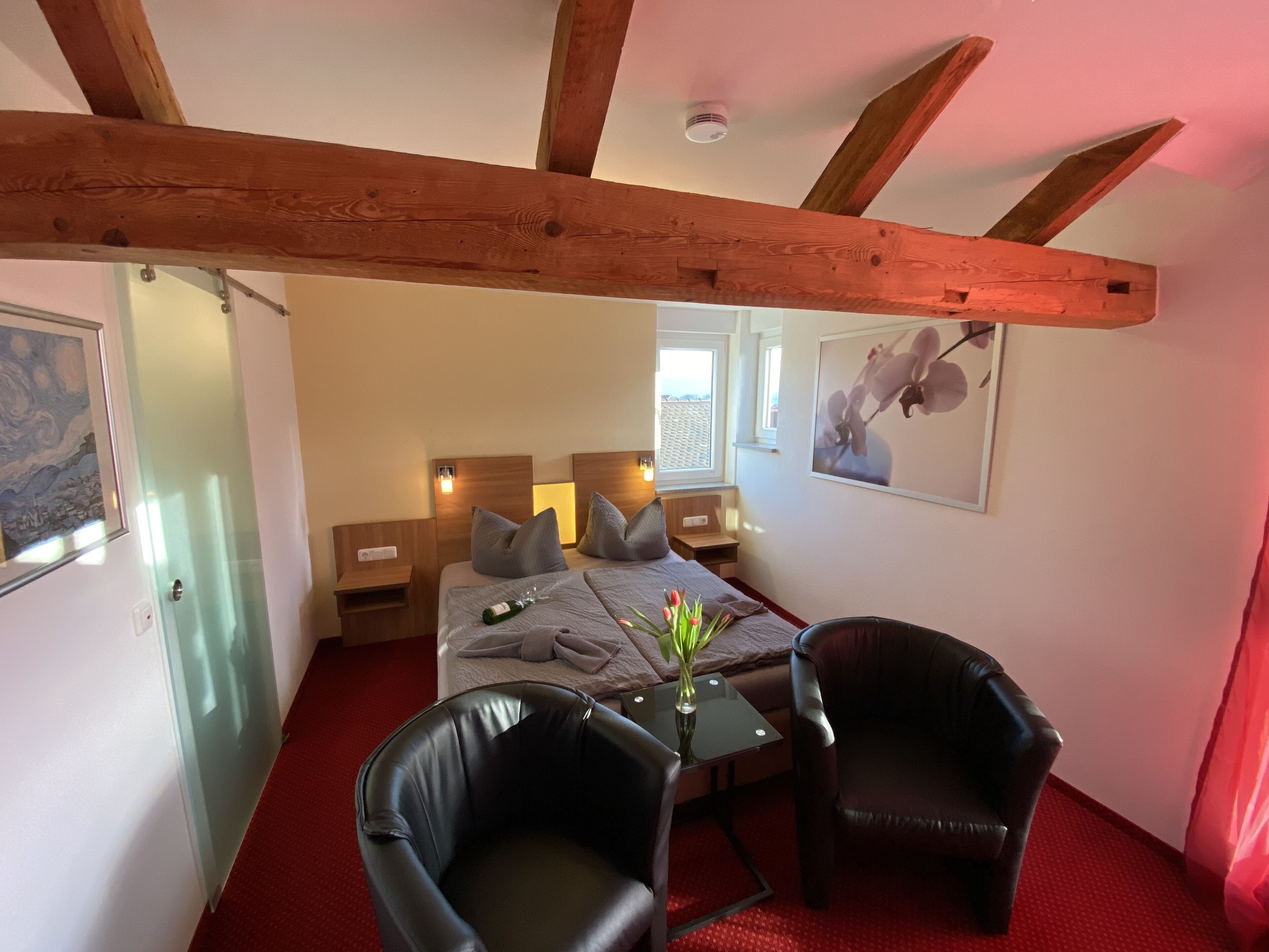 A hotel room with a bed, two nightstands, a glass table with a vase, two leather chairs, wooden beams on the ceiling, and artwork on the walls.