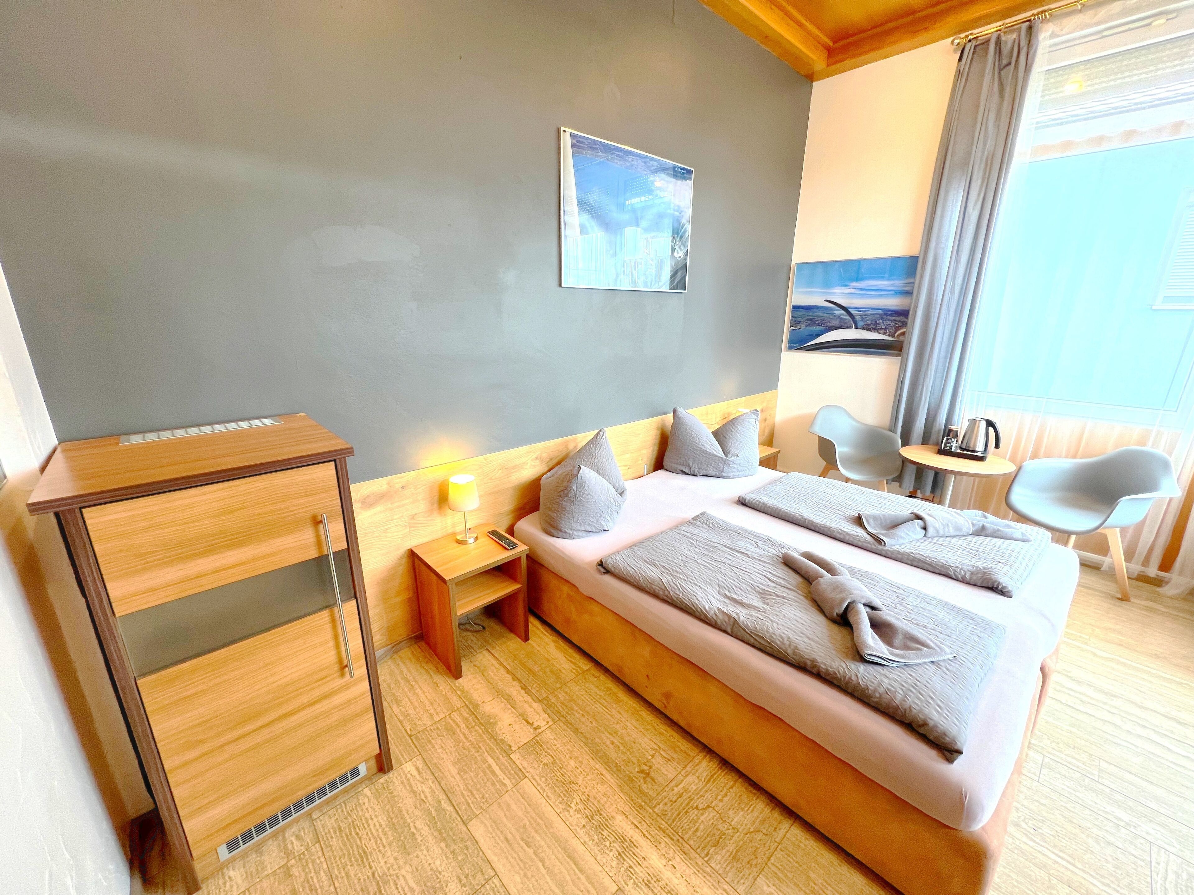 Habitación de hotel con cama, mesita de noche con lámpara, escritorio pequeño, silla y ventana con cortinas.