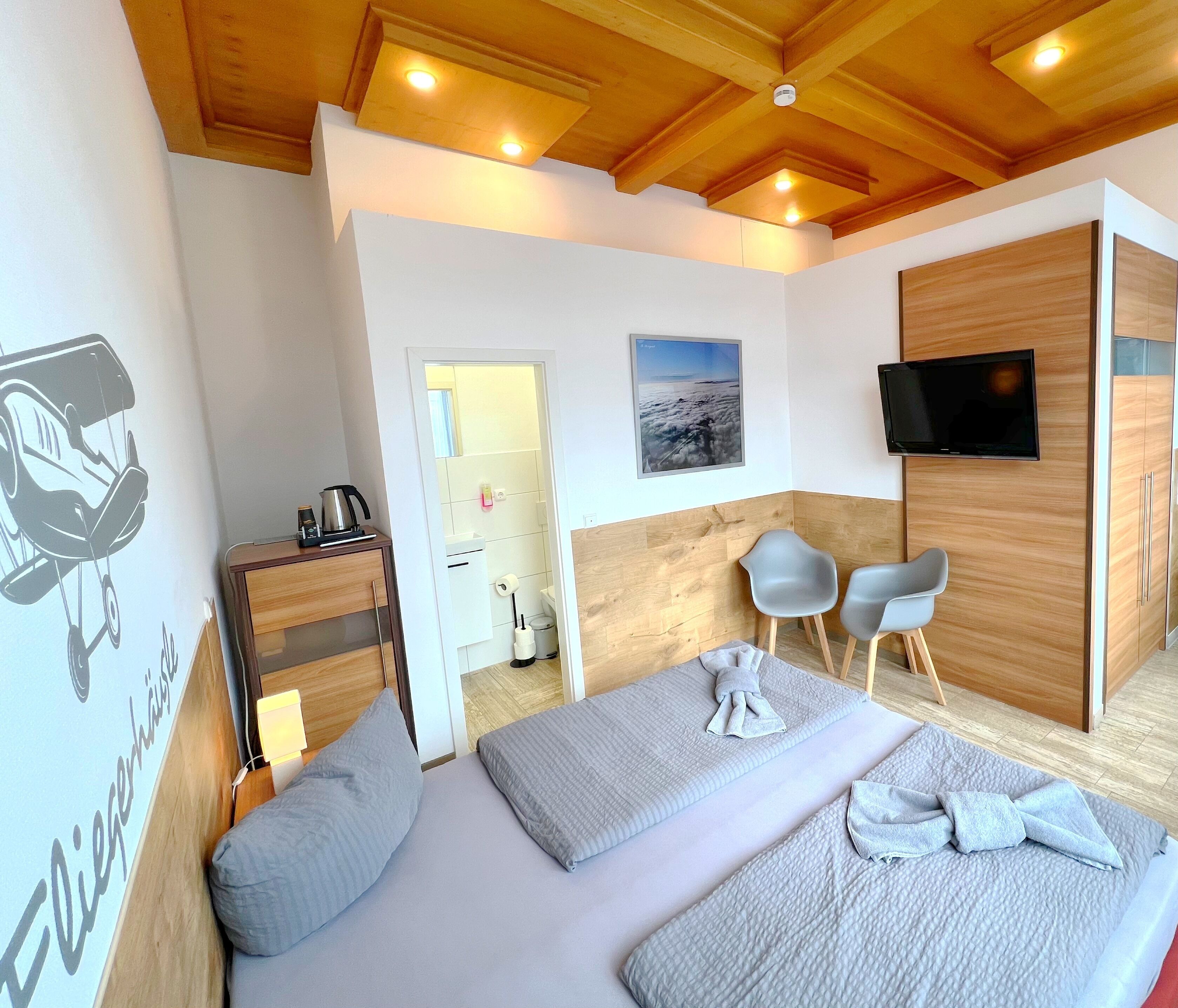 A hotel room with a wooden ceiling, a bed with grey bedding, a TV mounted on the wall, and a small seating area with two chairs.