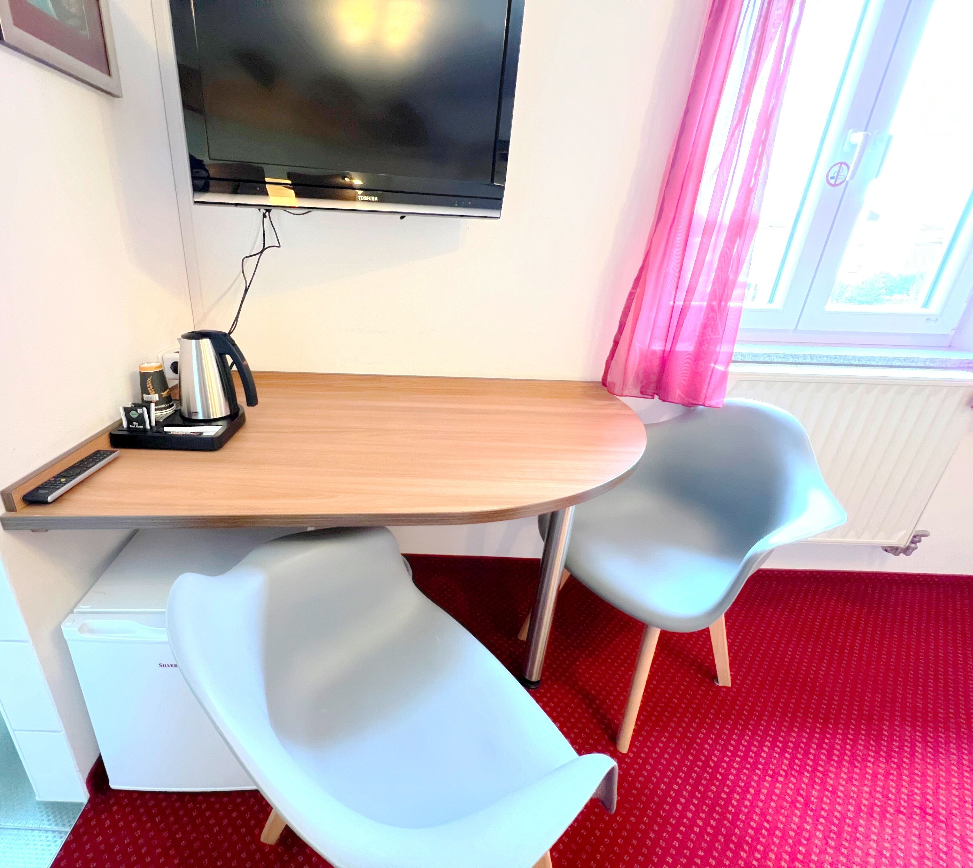 A small room with a wooden desk, two grey chairs, a television, and a radiator.