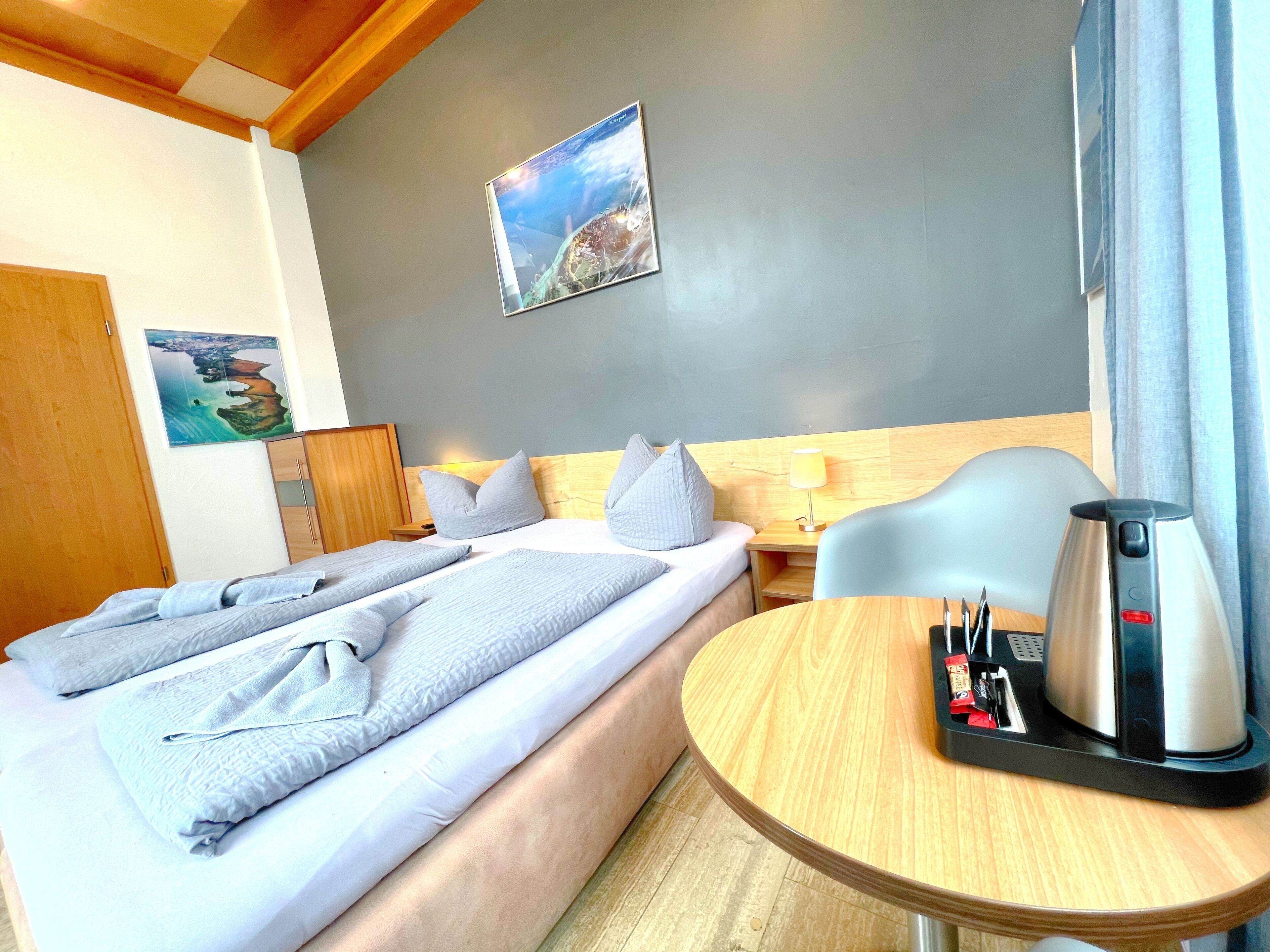 Habitación de hotel con cama, una mesita con hervidor, una silla, cabecera de madera y un cuadro en la pared.