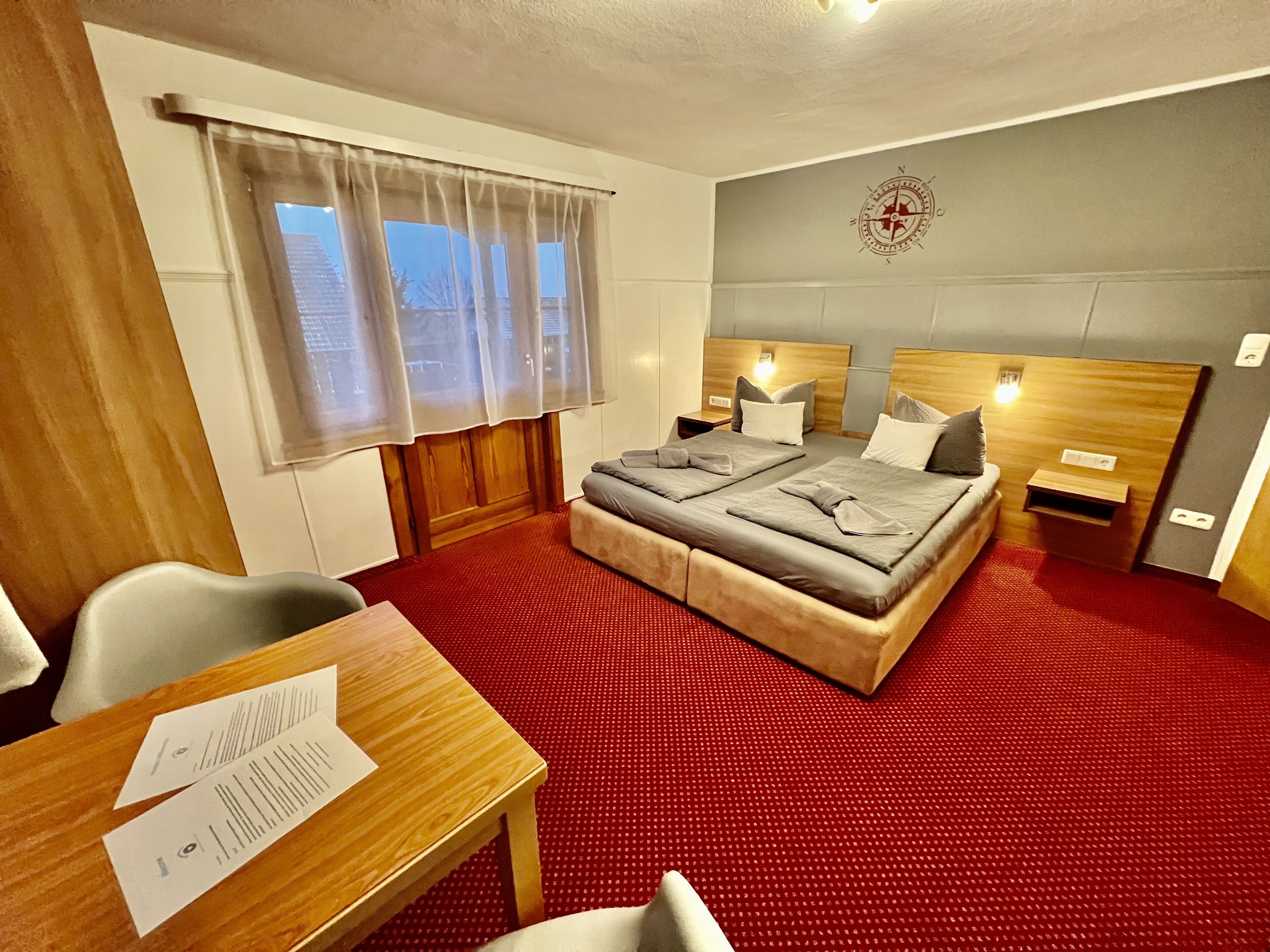 Une chambre d’hôtel avec un grand lit, deux tables de chevet, un bureau avec des papiers, une fenêtre avec des rideaux et une boussole fixée au mur.