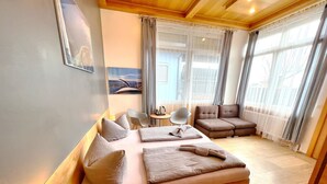 A modern hotel room with a large bed, a sofa, a small table, and a chair. There are two windows with curtains, a painting on the wall, and a radiator under the windows.