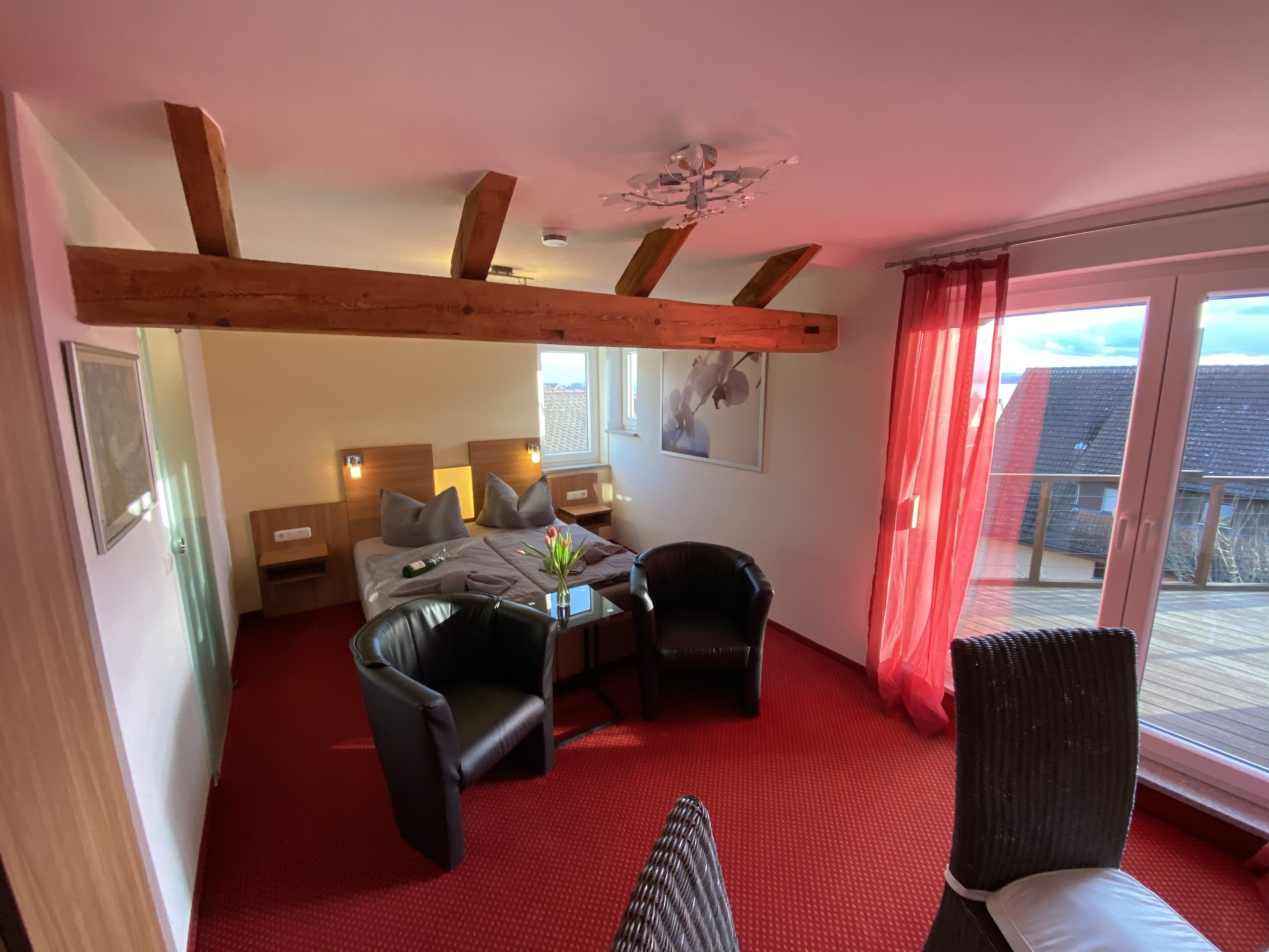 A hotel room with a bed, two chairs, a table with a vase, a ceiling fan, and a window with curtains.