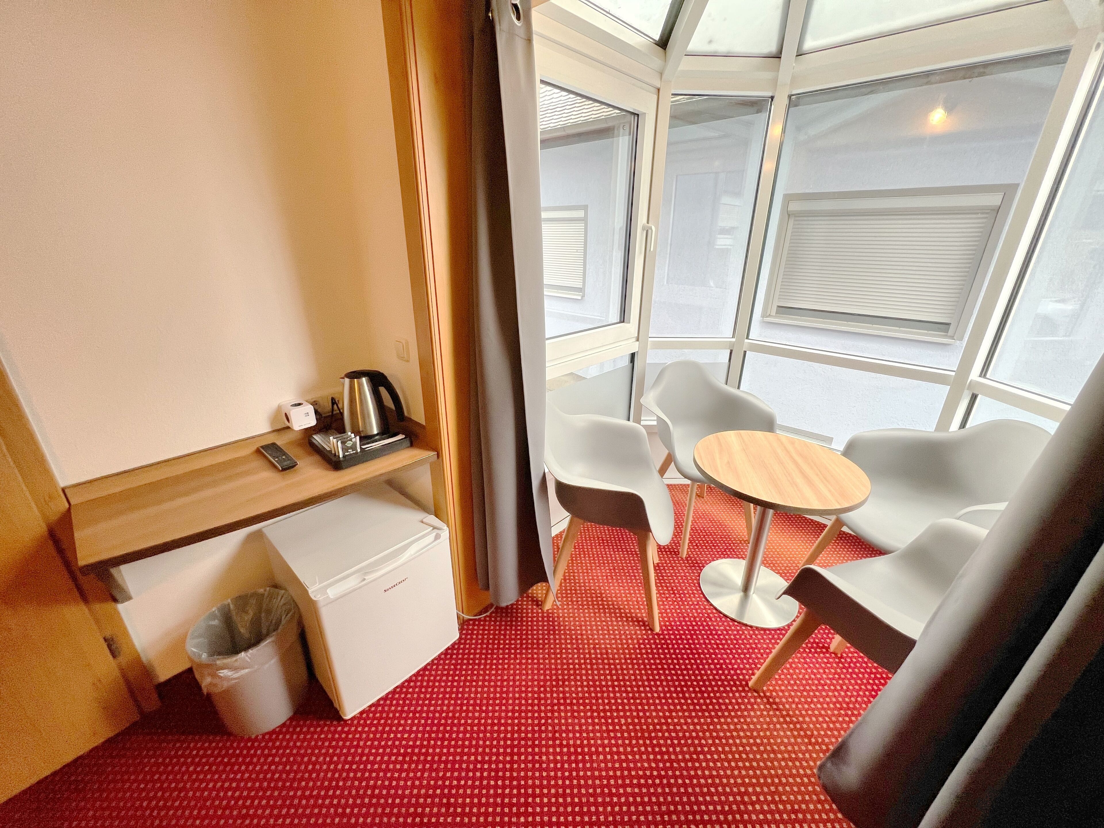 A compact room with a small table and two chairs, a mini fridge, a kettle, and a trash bin.