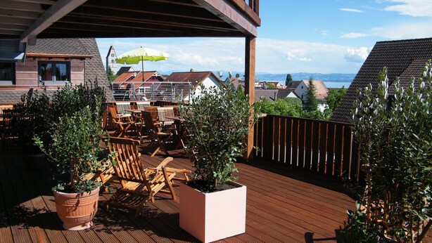 A wooden deck with outdoor seating, potted plants, and a view of houses and mountains in the background.