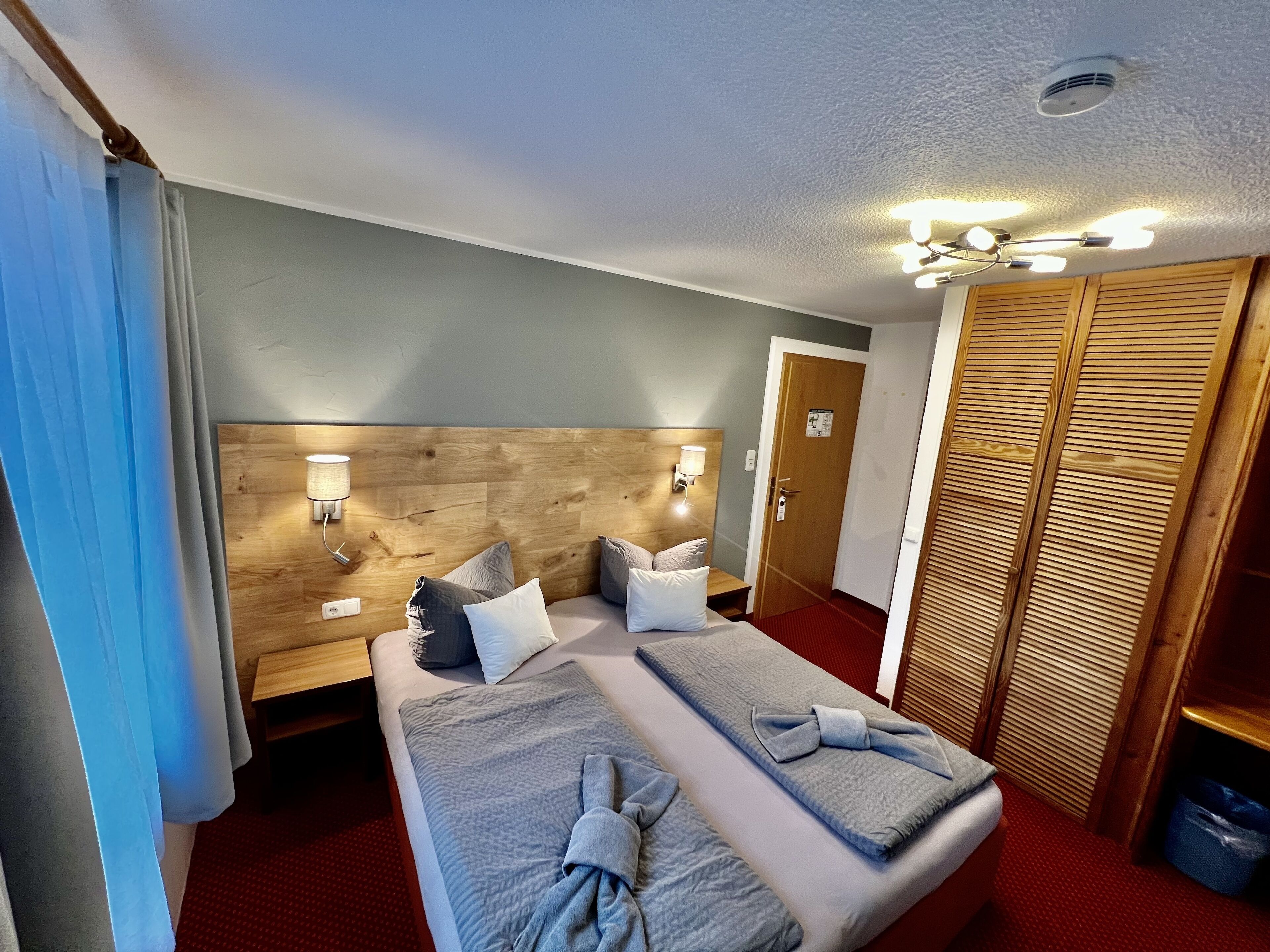 Habitación de hotel con dos camas, cabecero de madera, luces fijadas a la pared, una puerta y un armario.