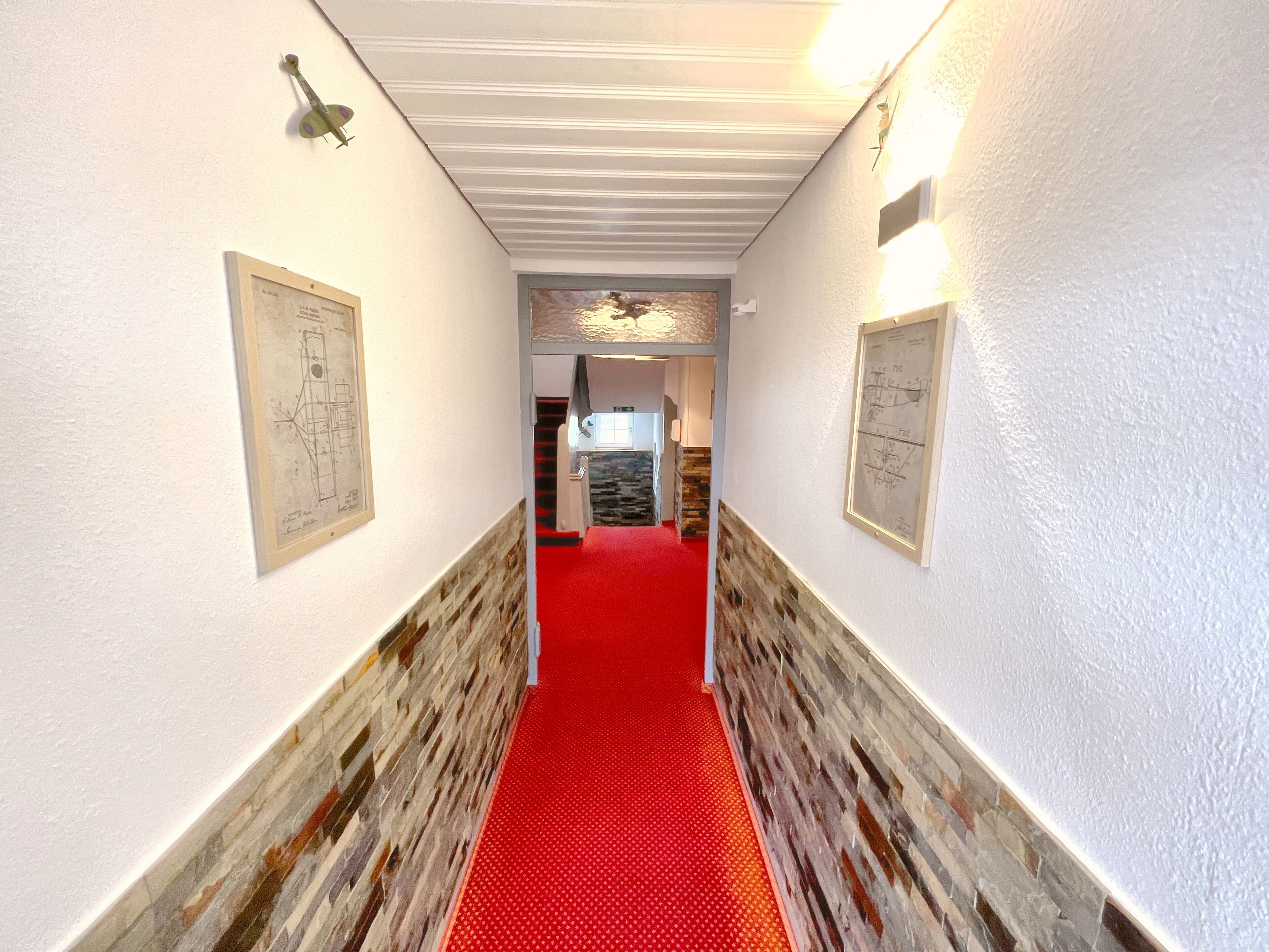 A hallway with a red carpet, white walls, and stone accents. There are framed architectural drawings on the walls.