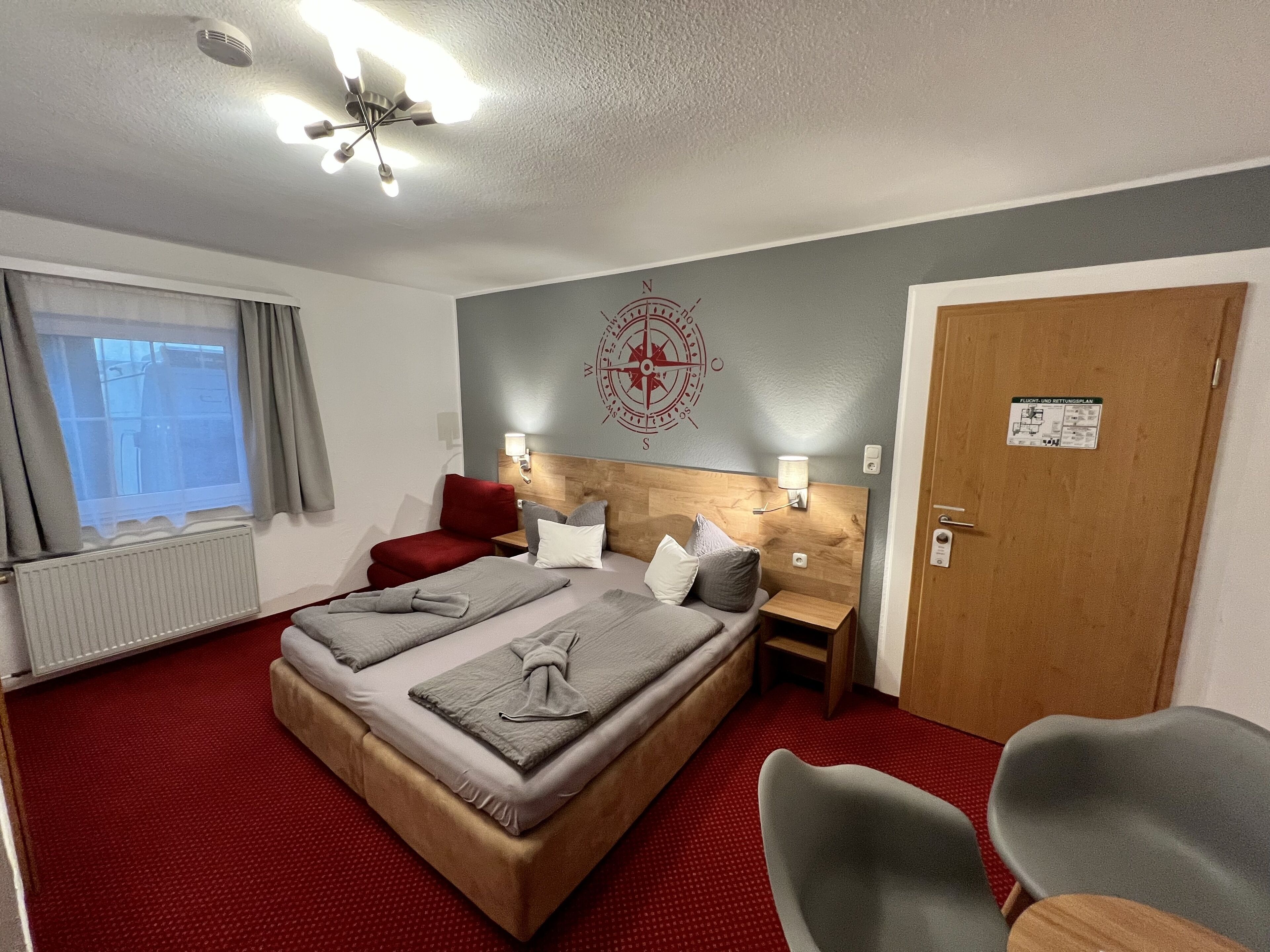 A hotel room with a large bed, two bedside tables, a red chair, a wooden door, and a window with curtains.