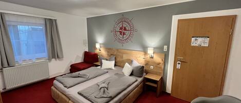 A hotel room with a large bed, two bedside tables, a red chair, a wooden door, and a window with curtains.