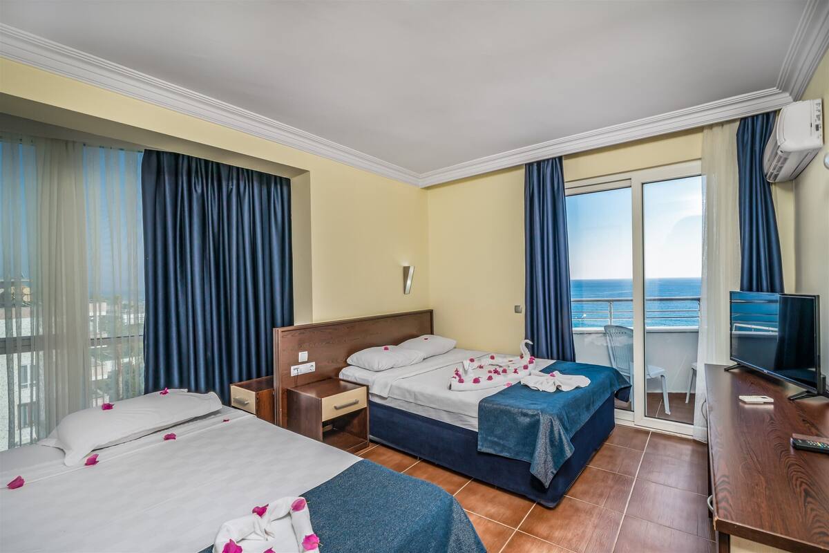 A hotel room with two beds, a TV, a desk, and a balcony with a view of the sea.