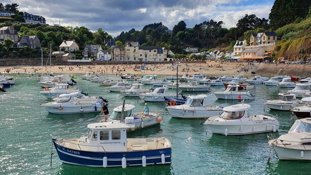 Uma marina costeira com vários barcos atracados, uma praia arenosa ao fundo e edifícios ao longo da costa.