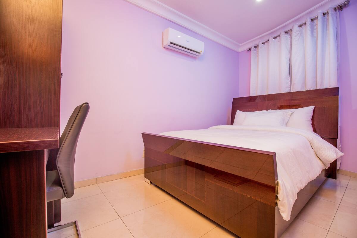 A bedroom with a bed, a desk, and an air conditioning unit.