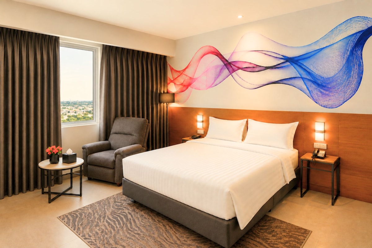 Een hotelkamer met een groot bed, een stoel, een nachtkastje en een muur met een kleurrijk abstract schilderij.