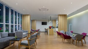 A modern hotel lobby with a wooden floor, grey sofas, and vibrant chairs.