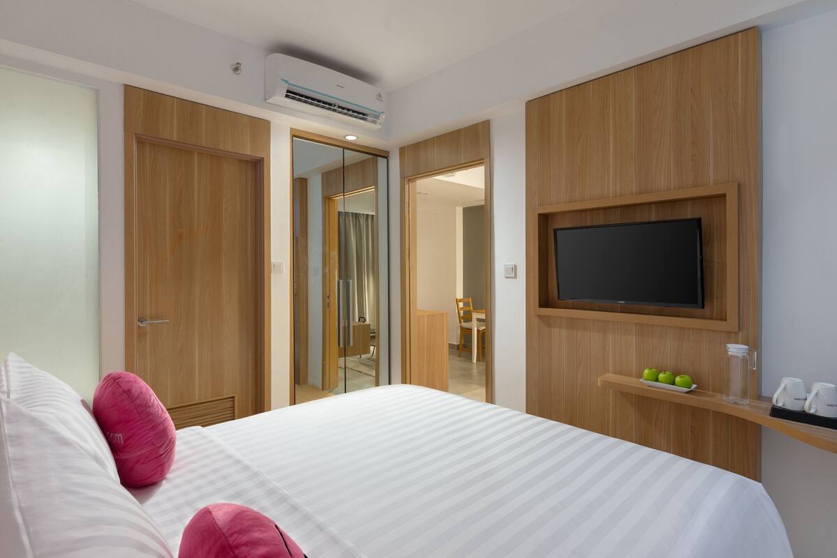 Habitación de hotel con cama, televisión, espejo y una pequeña estantería con objetos.