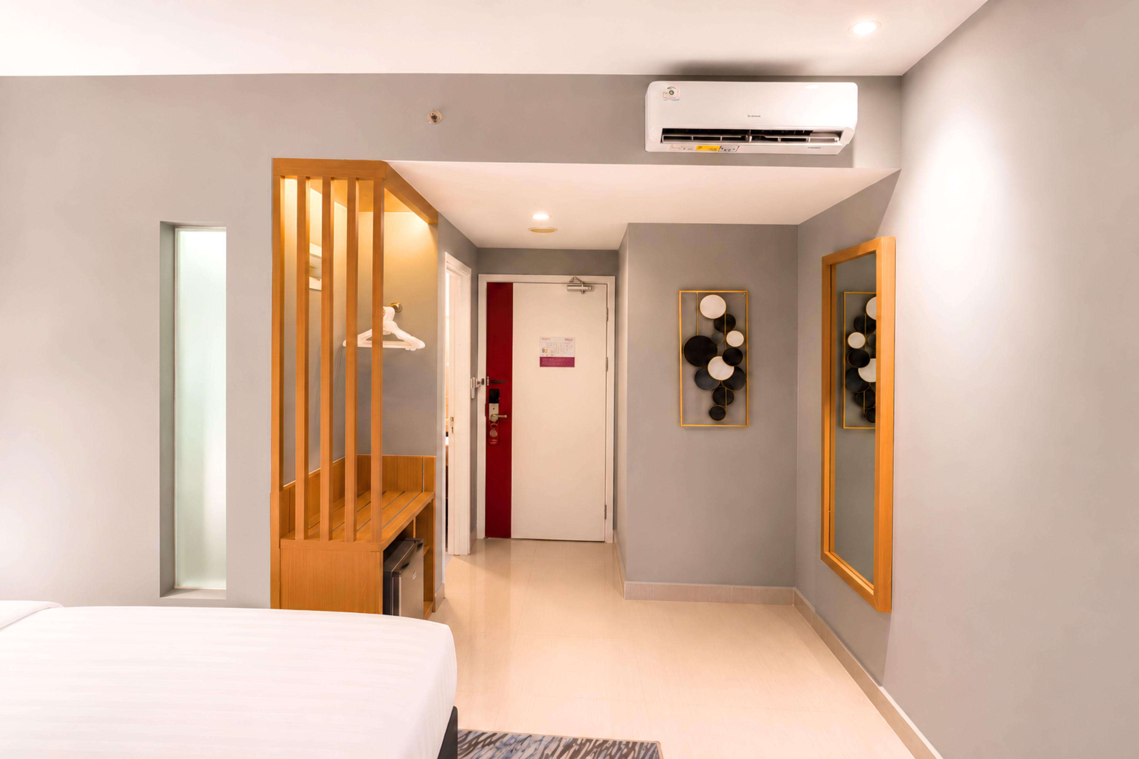 A modern hotel room with a bed, a wooden partition, an air conditioning unit, and a mirror with circular decorations.