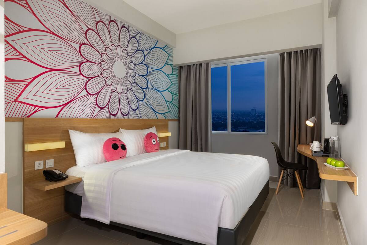 Una habitación de hotel moderna con un gran cuadro de flores en la pared, una cama con ropa blanca, un escritorio con una computadora y un plato de manzanas verdes, y vistas a la ciudad de noche.