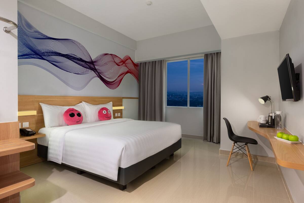 Habitación de hotel moderna con una cama grande, un escritorio con silla, televisión y vistas a la ciudad al anochecer.