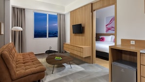 A modern hotel room with a sofa, a small table with fruit, a bed, a TV, and a view of the city at dusk.