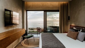 Habitación de hotel con una cama grande, televisión montada en la pared, vistas al mar y un balcón con sillas.