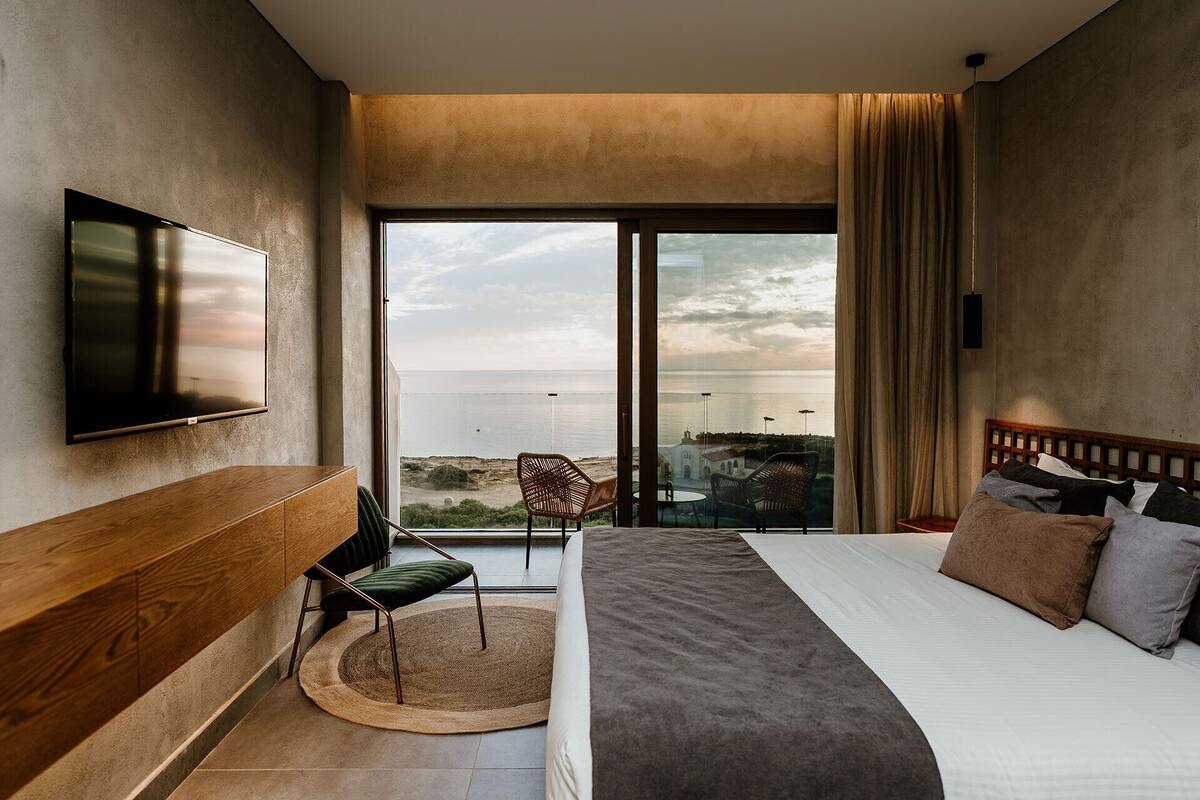 Ein Hotelzimmer mit einem großen Bett, einem an der Wand befestigten Fernseher, Meerblick und einem Balkon mit Sitzgelegenheiten.