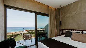 Habitación de hotel con un amplio ventanal corredera que da a un balcón con vista al mar y un pequeño edificio blanco.