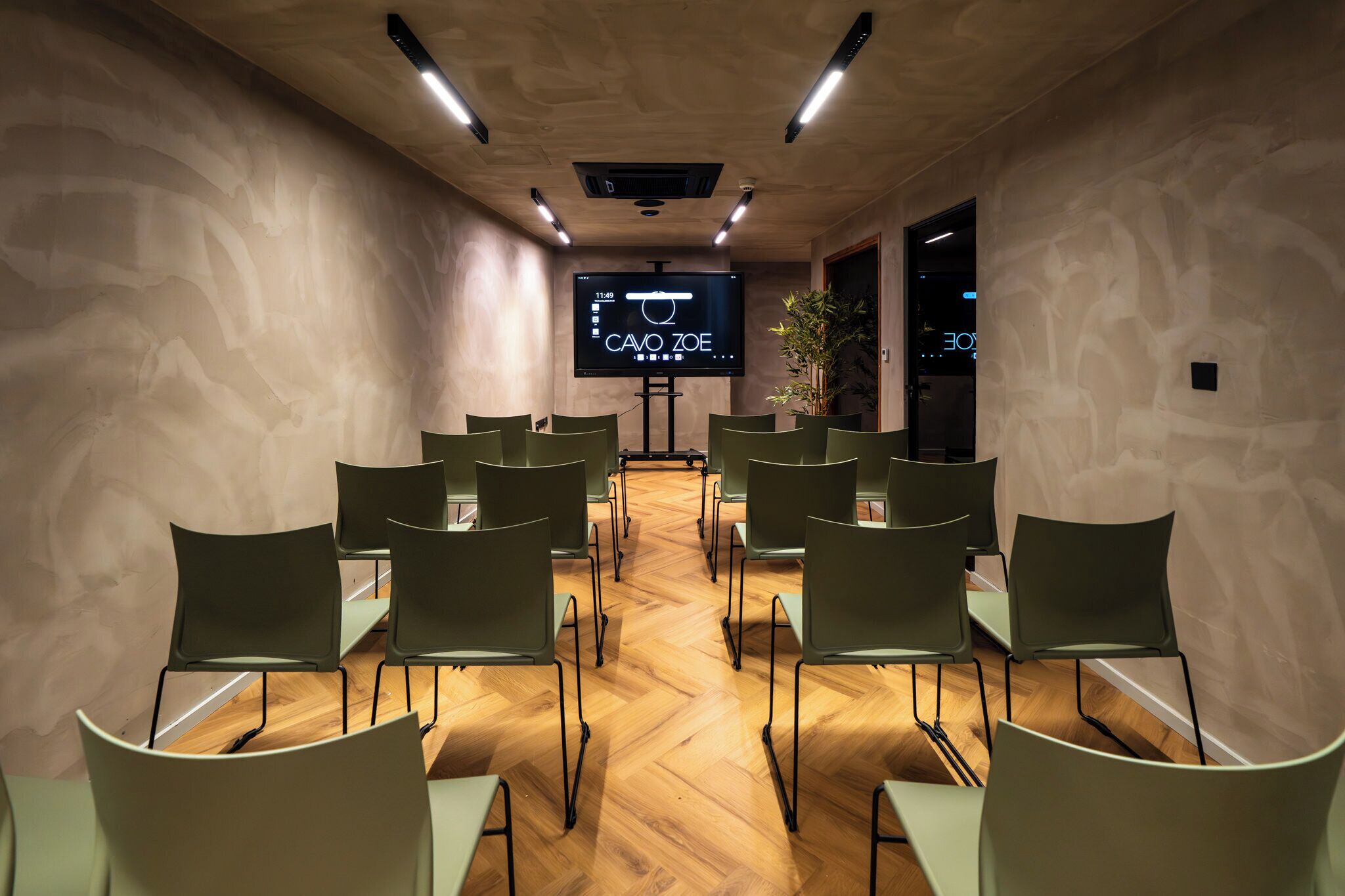 A conference room with a large screen, wooden floor, and green chairs arranged in rows.