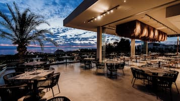 An outdoor dining area with tables and chairs, a palm tree, and a view of the sea at dusk.