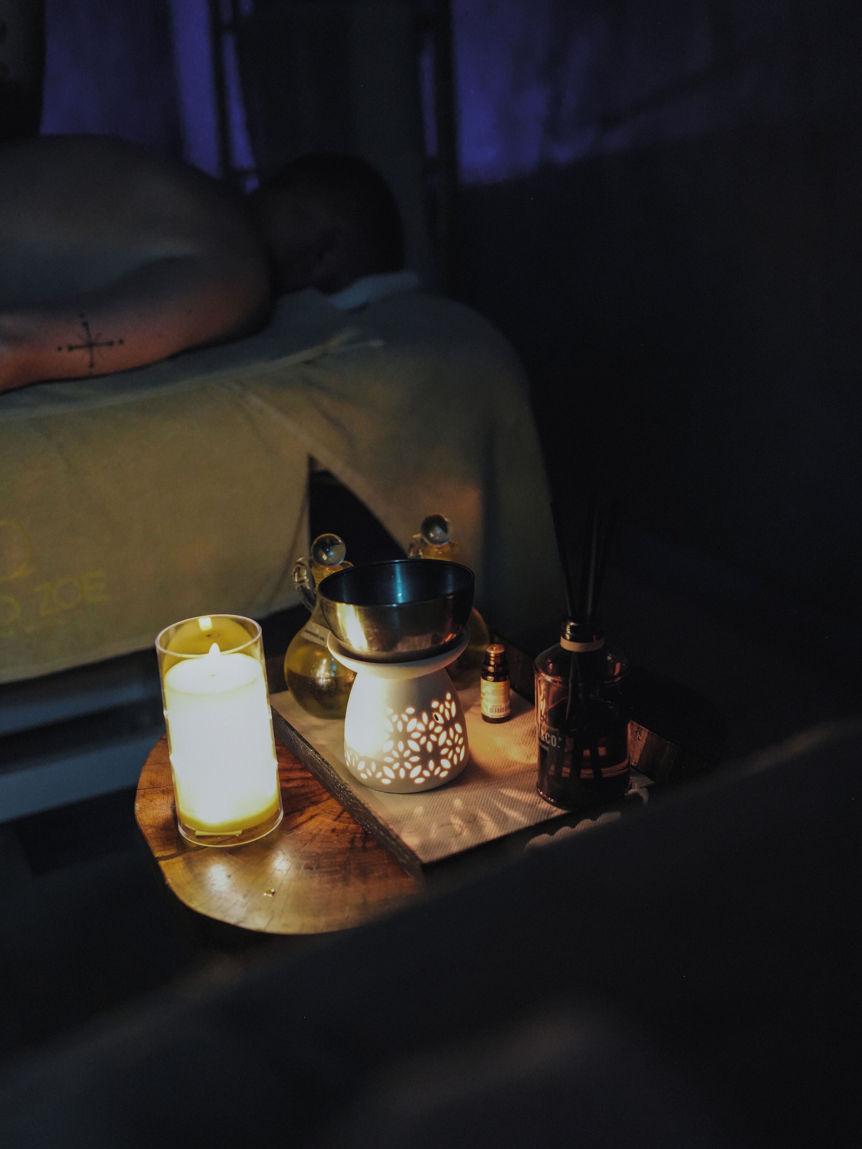 A dimly lit room with a bed, a candle, a lantern, and a bottle on a wooden tray.