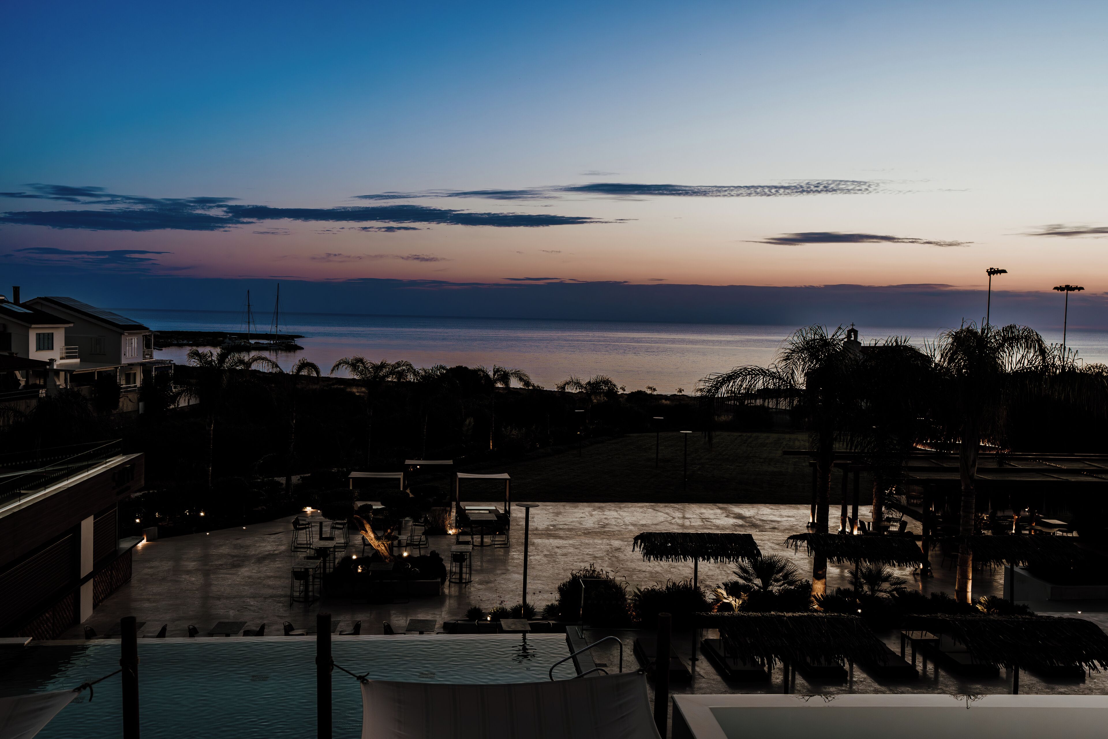 Zona de piscina con sillas de descanso y sombrillas, con vistas a una masa de agua y un cielo al atardecer.