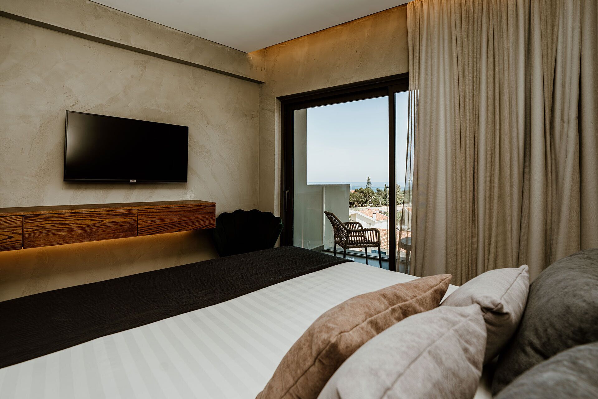 A modern hotel room with a large bed, a flat-screen TV mounted on the wall, and a balcony with a view of the sea.