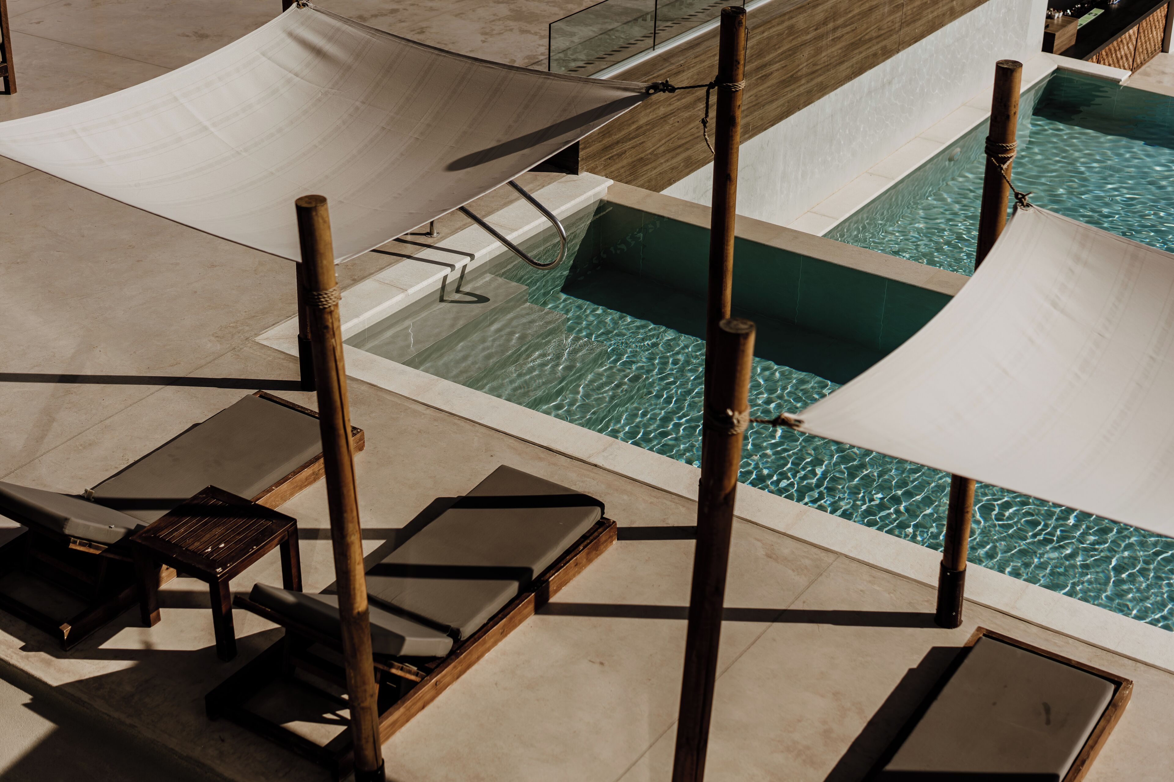 A pool area with multiple sun loungers and a small table, surrounded by a wooden deck and a bamboo pole structure.