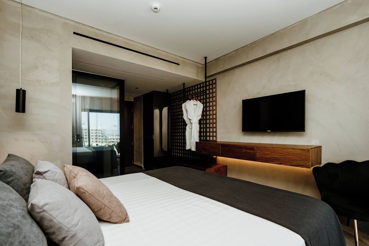 Ein modernes Hotelzimmer mit einem großen Bett, einem flachbildfernseher an der Wand, einem Holz-Kopfteil und Blick auf die Stadt durch das Fenster.