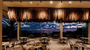 Un área de comedor al aire libre con mesas y sillas, ventanas grandes y vistas al cielo.