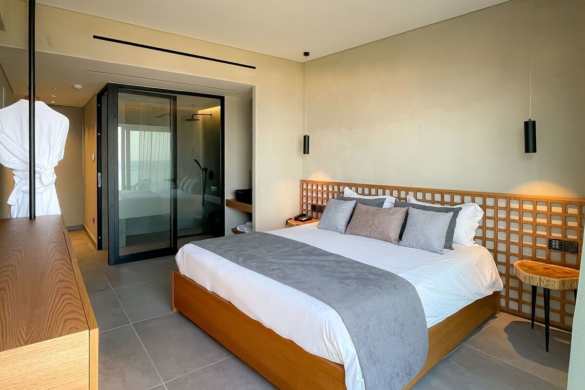 A hotel room with a bed, a wooden dresser, a robe rack, a glass door leading to a balcony, and a nightstand with a lamp.
