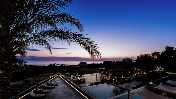 Zona de piscina en un resort con sillones de descanso y vista al atardecer sobre el agua.